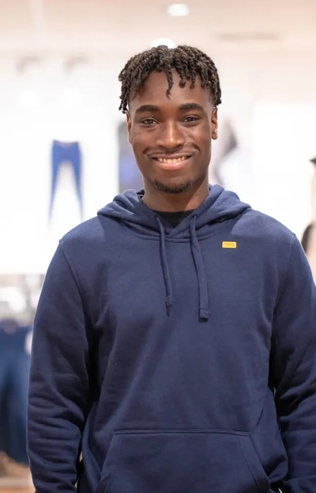Ein junger Mann mit kurzen, geflochtenen Haaren lächelt in die Kamera. Er trägt einen dunkelblauen Kapuzenpullover mit einem kleinen gelben Etikett und steht in einem Innenbereich vor einem unscharfen Hintergrund.