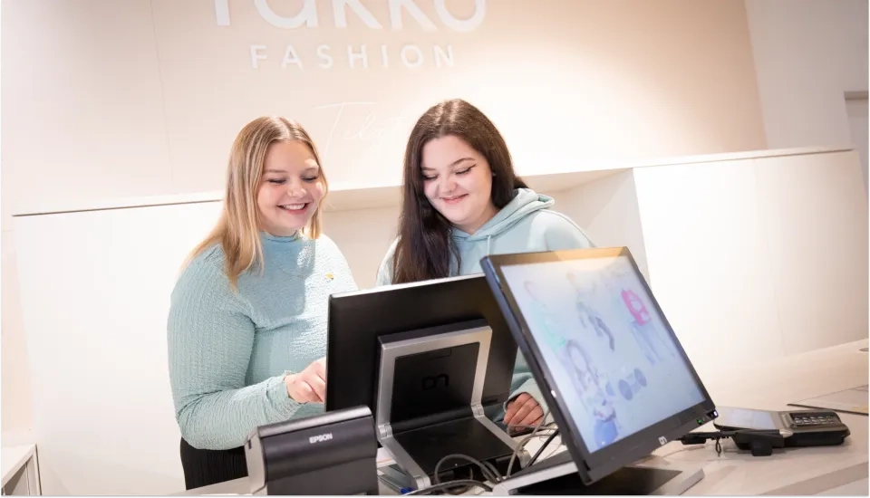 Zwei Frauen stehen lächelnd hinter einer Ladentheke und schauen auf einen Computerbildschirm. Sie scheinen in einem Bekleidungsgeschäft zu arbeiten. An der Wand hinter ihnen ist Fakko Fashion zu sehen.