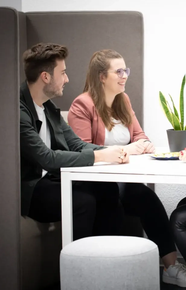 Zwei Personen sitzen lächelnd in einer modernen Bürokabine an einem weißen Tisch mit einer Topfpflanze und Tellern zusammen und unterhalten sich. Die Atmosphäre wirkt ungezwungen und kollegial.
