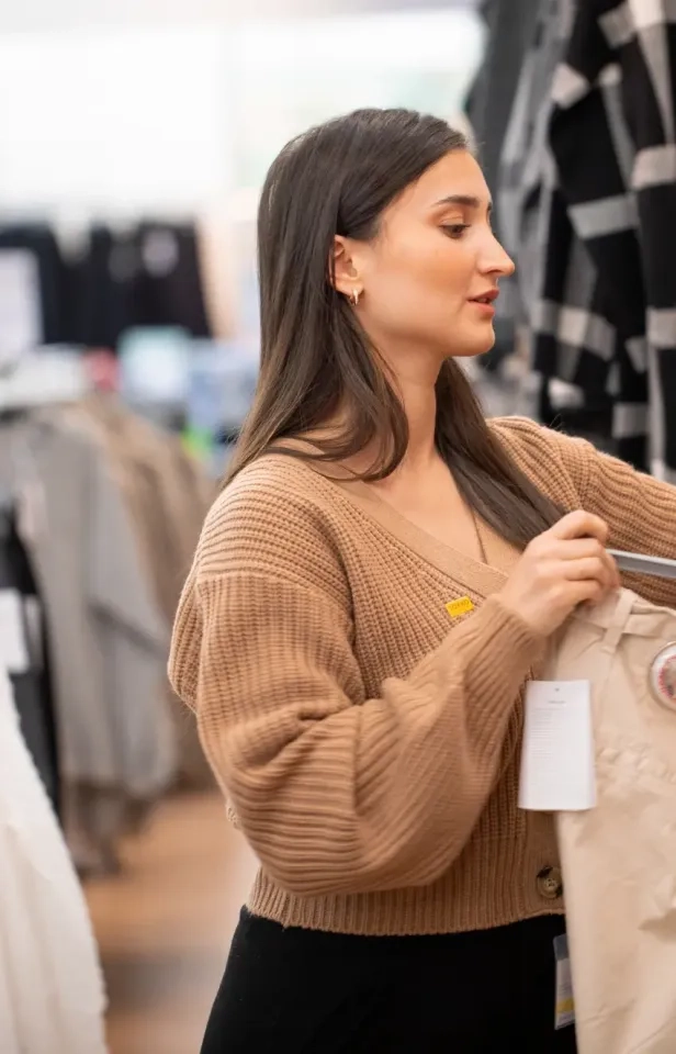 Eine Frau mit langen braunen Haaren und einem hellbraunen Pullover hält beim Kleiderkauf in einem Geschäft ein beiges Kleidungsstück hoch. Sie betrachtet den Artikel aufmerksam.