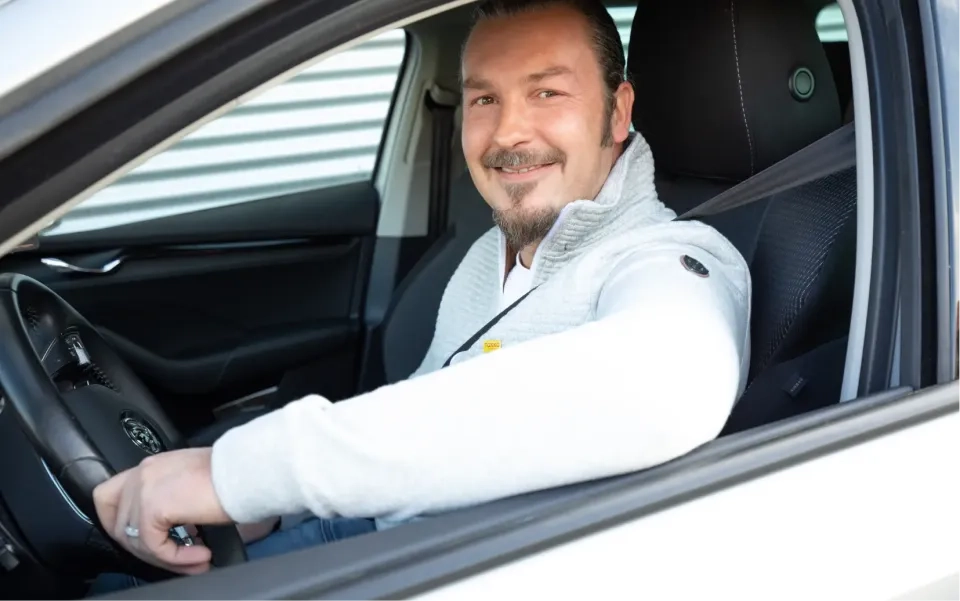 Ein lächelnder Mann mit Spitzbart sitzt auf dem Fahrersitz eines Autos. Er trägt eine hellgraue Jacke und einen Sicherheitsgurt und blickt durch das offene Fenster in die Kamera.