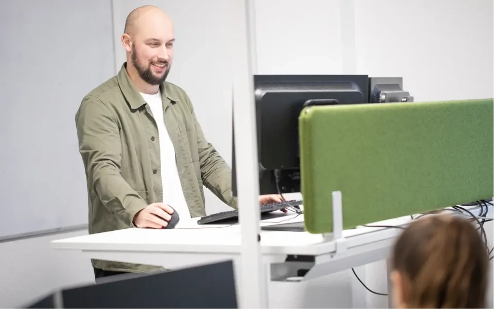Ein Mann mit Bart und grüner Jacke steht lächelnd an einem weißen Schreibtisch und bedient in einer modernen Büroumgebung eine Computermaus und eine Tastatur.