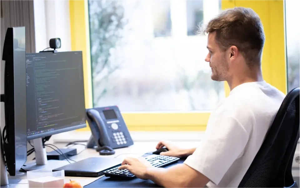 Ein Mann sitzt an einem Schreibtisch und arbeitet an einem Computer, auf dessen Monitor Code angezeigt wird. Er benutzt Tastatur und Maus. Im Hintergrund sind ein Telefon, eine Webcam und ein Fenster zu sehen.