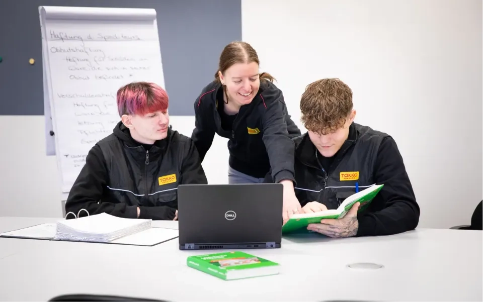 Drei Personen in schwarzen Uniformen sitzen an einem Tisch. Eine Person steht lächelnd da und zeigt auf einen grünen Ordner, den eine sitzende Person hält. Eine andere Person beobachtet. Auf dem Tisch liegen ein Laptop, ein Buch und ein Flipchart mit Notizen.