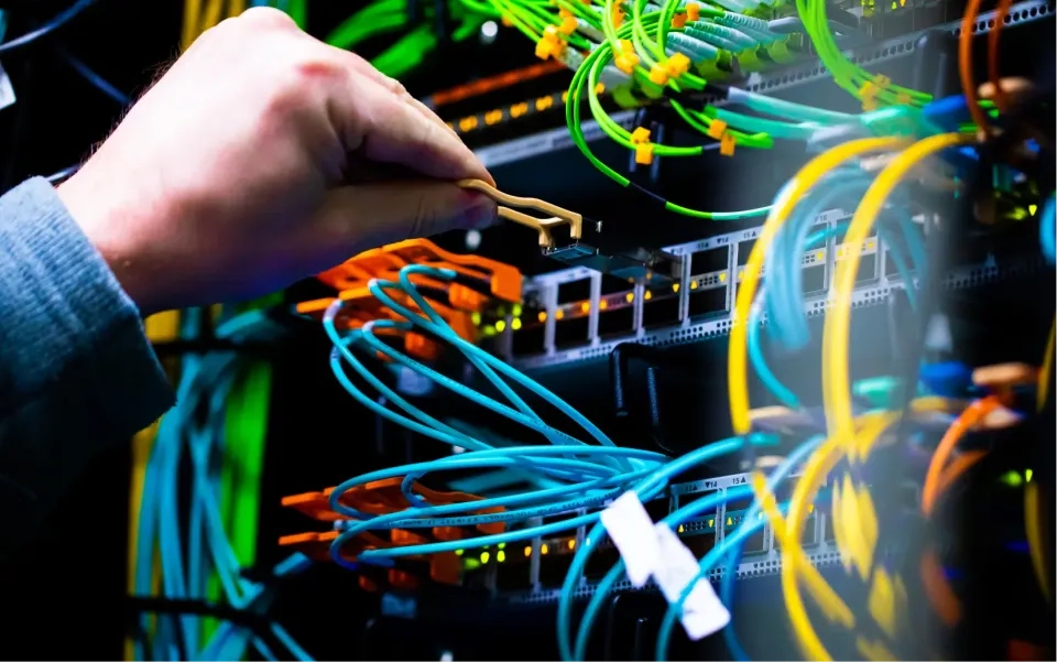 Die Hand einer Person richtet ein Netzwerkkabel in einem Server-Rack eines Rechenzentrums aus, das voller bunter angeschlossener Kabel und blinkender Anzeigeleuchten ist.