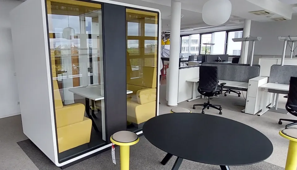 Ein moderner Büroraum mit Schreibtischen und Stühlen, darunter eine Telefonzelle mit Glaswänden und gelben Stühlen, ein runder schwarzer Tisch und ein kleiner gelber Hocker im Vordergrund. Große Fenster sorgen für Tageslicht.