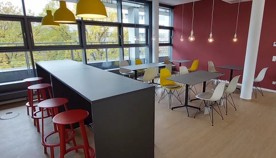 Eine moderne Cafeteria mit großen Fenstern, roten und gelben Stühlen, roten Barhockern, schwarzen Tischen und hängenden Pendelleuchten, mit einer roten Akzentwand und Holzboden.