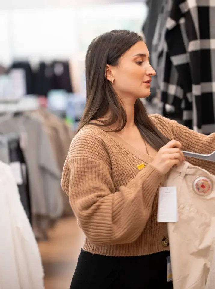 Eine Frau mit langen dunklen Haaren und einem braunen Pullover betrachtet in einem Bekleidungsgeschäft eine beige Hose auf einem Kleiderbügel. Im Hintergrund sind weitere Kleidungsstücke und Kleiderständer zu sehen.