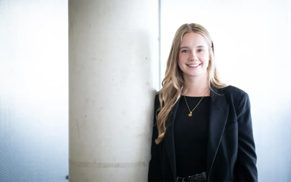 Eine junge Frau mit langen blonden Haaren, einem schwarzen Blazer und einer goldenen Halskette steht in einem Gebäude neben einer großen Betonsäule und lächelt vor einem hellen, sanft beleuchteten Hintergrund in die Kamera.