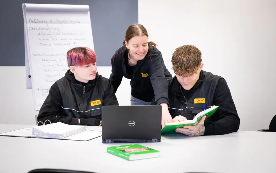 Drei junge Leute in schwarzen TOKS-Uniformen arbeiten gemeinsam an einem Tisch mit Laptop und Ordnern. Einer steht lächelnd da und zeigt auf ein Dokument, während die anderen sitzen und es aufmerksam betrachten.