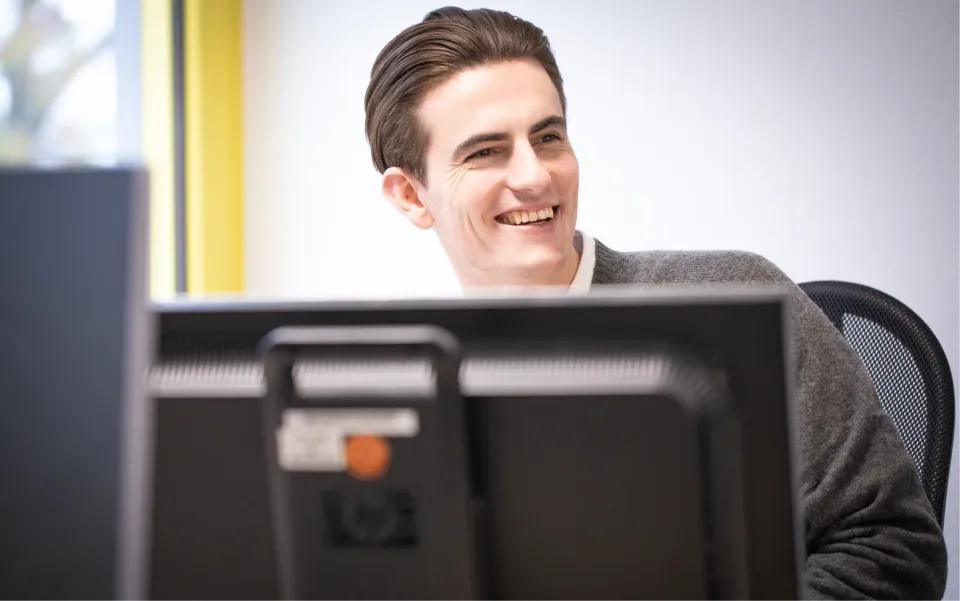 Ein Mann mit dunklem Haar und einem grauen Pullover sitzt lächelnd an einem Schreibtisch hinter zwei Computermonitoren in einem hell erleuchteten Büro.