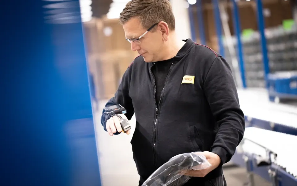 Ein Mann mit Schutzbrille und schwarzer Jacke scannt in einem Lagerhaus einen verpackten Artikel mit einem tragbaren Barcode-Scanner. Im Hintergrund sind Regale und Kisten zu sehen.