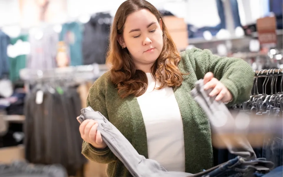 Eine Frau mit langen braunen Haaren und einem grünen Pullover betrachtet beim Einkaufen in einem Bekleidungsgeschäft eine graue Jeans. Im Hintergrund sind Kleiderständer mit Kleidung zu sehen.