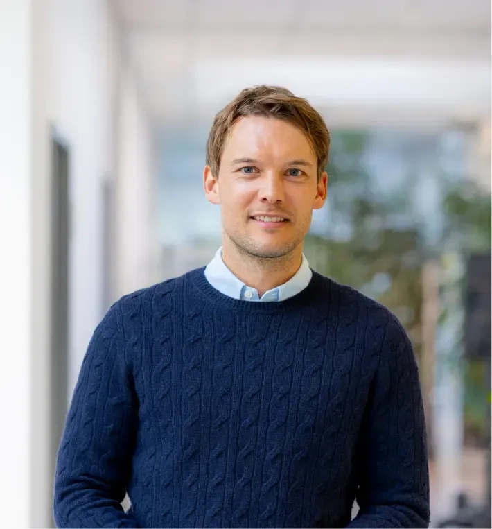 Ein Mann mit hellbraunem Haar, der einen marineblauen Zopfmusterpullover über einem hellblauen Hemd trägt, steht in einem hellen Flur mit verschwommenem Grün im Hintergrund.
