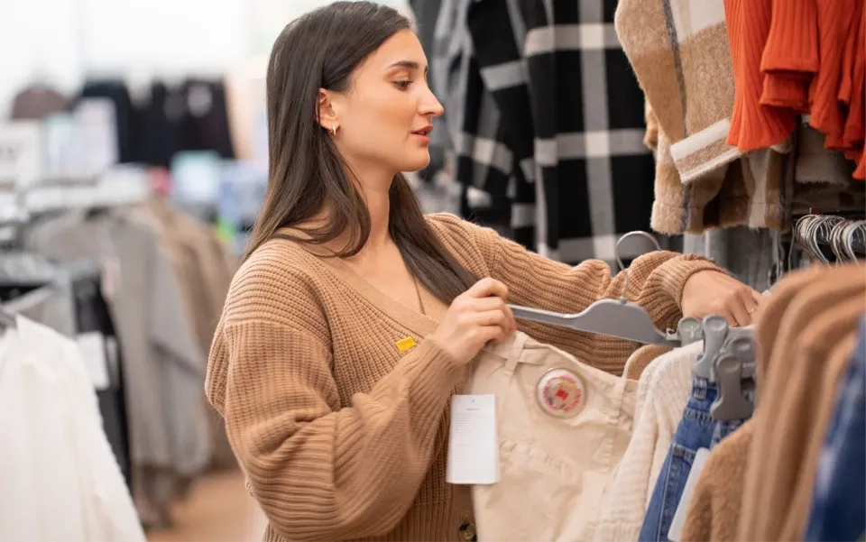 Eine Frau mit langen dunklen Haaren stöbert in einem Geschäft nach Kleidung. Sie hält beige Hosen auf einem Kleiderbügel und betrachtet Pullover auf einer Stange. Sie trägt einen braunen Pullover und scheint sich auf den Einkauf zu konzentrieren.