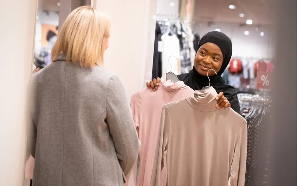 Eine Frau mit Hijab lächelt, während sie in einem Bekleidungsgeschäft zwei Rollkragenpullover hochhält und sie einer anderen Frau mit blonden Haaren und einem grauen Mantel zeigt.