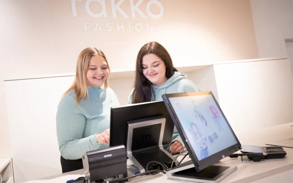 Zwei junge Frauen stehen lächelnd hinter einer Kasse in einem Takko Fashion Store und bedienen eine Touchscreen-Kasse. Auf dem Tresen sind Computermonitore, ein Belegdrucker und ein Bezahlterminal zu sehen.