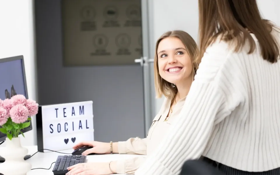 Zwei Frauen in einem Büro. Eine sitzt an einem Schreibtisch mit Computer und lächelt die andere an, die steht. Auf dem Schreibtisch steht ein Schild mit der Aufschrift „TEAM SOCIAL“ und Herzen. Daneben steht eine Vase mit rosa Blumen.