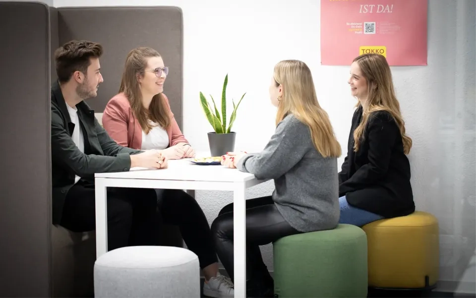 Vier junge Erwachsene sitzen in einem modernen Büro um einen weißen Tisch und unterhalten sich. Auf dem Tisch steht eine Topfpflanze, an der Wand dahinter hängt ein rosa Poster.