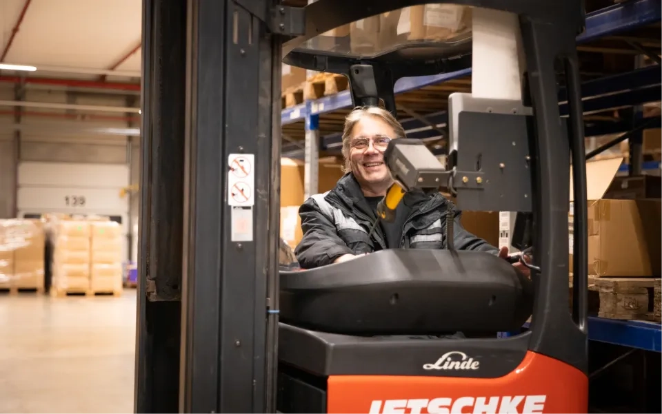 Ein lächelnder Arbeiter mit Brille und Jacke bedient einen Linde-Gabelstapler in einem Lager voller Regale und Kisten.