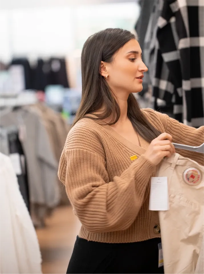 Eine Frau mit langen braunen Haaren und einem hellbraunen Strickpullover kauft in einem Geschäft Kleidung. Sie hält eine helle Hose auf einem Kleiderbügel und betrachtet sie genau. Im Hintergrund stehen Kleiderständer.