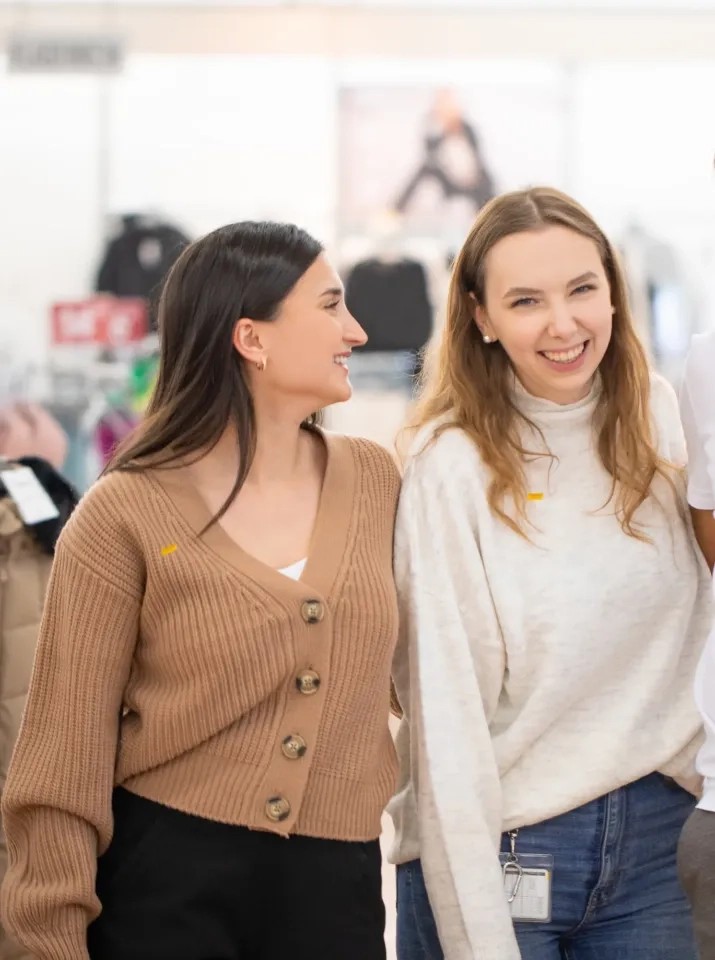Zwei Frauen stehen dicht beieinander in einem Raum, lächeln und wirken glücklich. Die eine trägt eine braune Strickjacke und blickt die andere an, die einen weißen Pullover trägt und ein Schlüsselband um den Hals hat. Im Hintergrund scheint ein Geschäft zu sein.