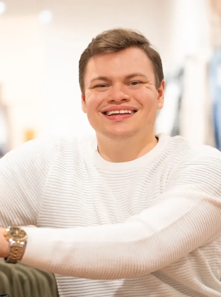 Ein Mann in einem weißen Rippenpullover und einer goldenen Uhr lächelt herzlich in die Kamera und sitzt in einem sanft beleuchteten Raum.