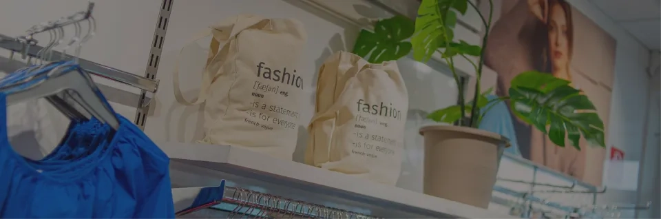 Regaldisplay mit blauer Kleidung, zwei beigen Tragetaschen mit der Aufschrift „Mode“, einer Topfpflanze und einem großen Frauenfoto im Hintergrund.