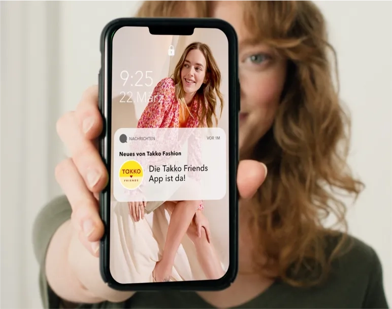 Eine Person hält ein Smartphone hoch. Auf dem Sperrbildschirm ist eine lächelnde Frau in Pink zu sehen. Auf Deutsch gibt es eine Benachrichtigung über die neue Takko Friends App. Das Datum ist der 22. März, 9:25 Uhr.