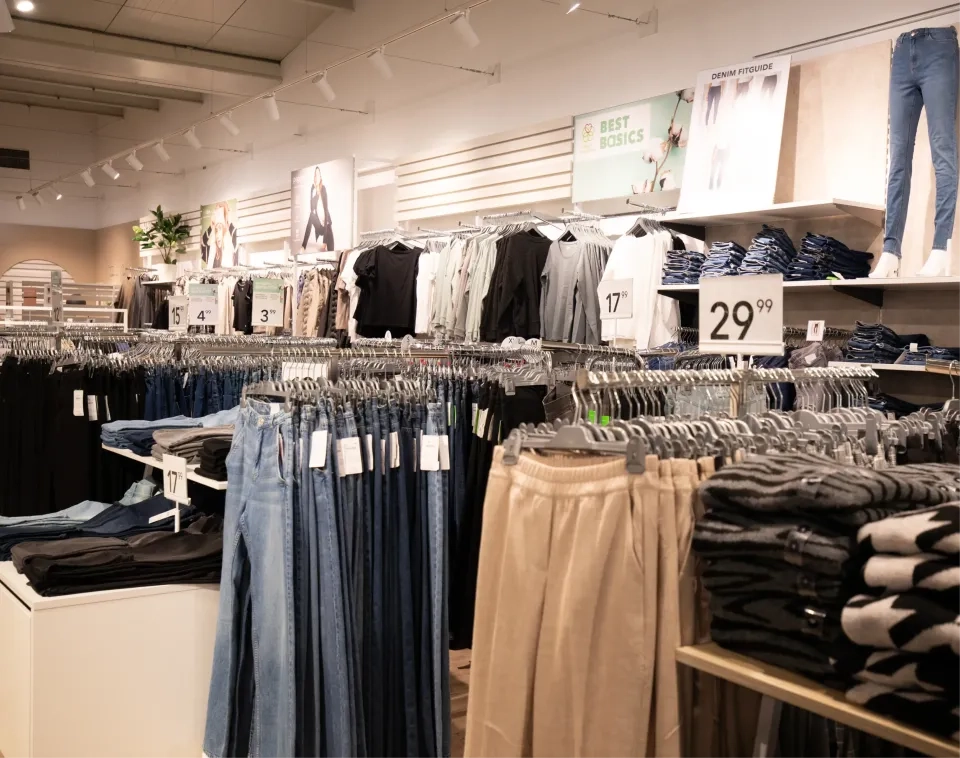 Innenansicht eines Bekleidungsgeschäfts mit Kleiderständern voller Jeans, Hosen, Hemden und Pullovern. Preisschilder sind sichtbar, und in den Regalen liegen gefaltete Kleidungsstücke. Schilder darüber werben für Basics und Denim-Styles. Helle Deckenbeleuchtung erhellt den Raum.