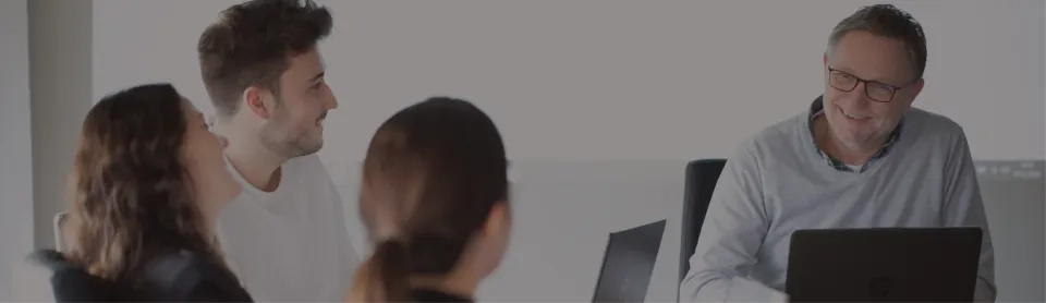 Vier Personen sitzen mit Laptops um einen Tisch und unterhalten sich. Ein Mann mit Brille lächelt, während die anderen ihm aufmerksam zuhören. Die Szene scheint eine Besprechung oder ein gemeinsamer Arbeitsbereich zu sein.