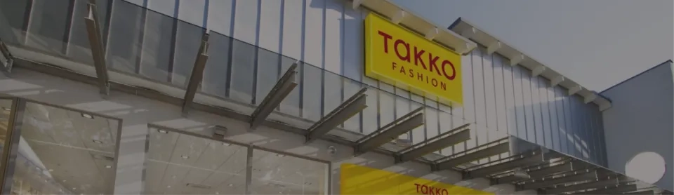 Schaufenster eines TOKKO Fashion-Einzelhandelsgeschäfts mit einem gelben Schild mit roter Schrift über dem Eingang und großen Fenstern, in denen sich der Himmel spiegelt.