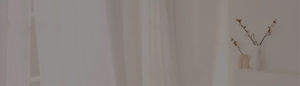 Minimalistisches Interieur mit transparenten weißen Vorhängen und sanfter Beleuchtung. Rechts steht auf einem kleinen weißen Regal eine beige Vase mit zarten Zweigen, an denen kleine weiße und braune Blüten hängen. Das schafft eine ruhige, heitere Atmosphäre.