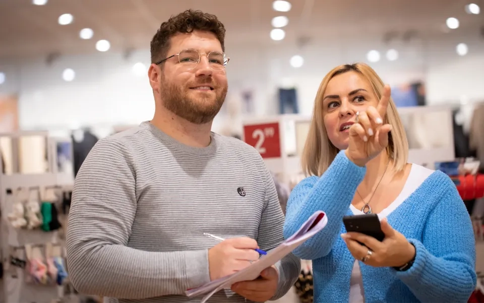 Zwei Menschen stehen in einem Geschäft. Der Mann hält lächelnd einen Notizblock und einen Stift in der Hand, während die Frau nach vorne zeigt und ein Telefon in der Hand hält. Beide scheinen zwischen den Warenregalen in ein Gespräch vertieft zu sein.