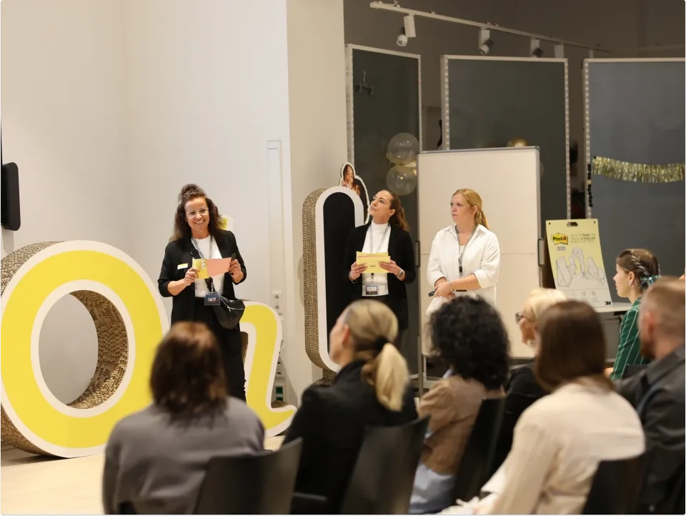 Drei Frauen stehen vor einem modernen Raum und präsentieren vor einem sitzenden Publikum. Im Hintergrund sind große gelbe und weiße Buchstaben, Poster, Luftballons und eine weiße Tafel zu sehen.