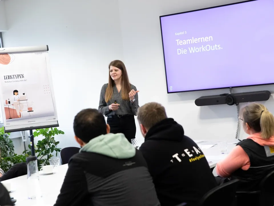 Eine Frau steht vor einer Gruppe und präsentiert in einem hellen Raum. Hinter ihr zeigt ein Bildschirm „Teamlernen Die Workouts“. Außerdem ist ein Flipchart mit der Überschrift „Lerntypen“ zu sehen. Die Anwesenden sitzen an einem Tisch und hören aufmerksam zu.