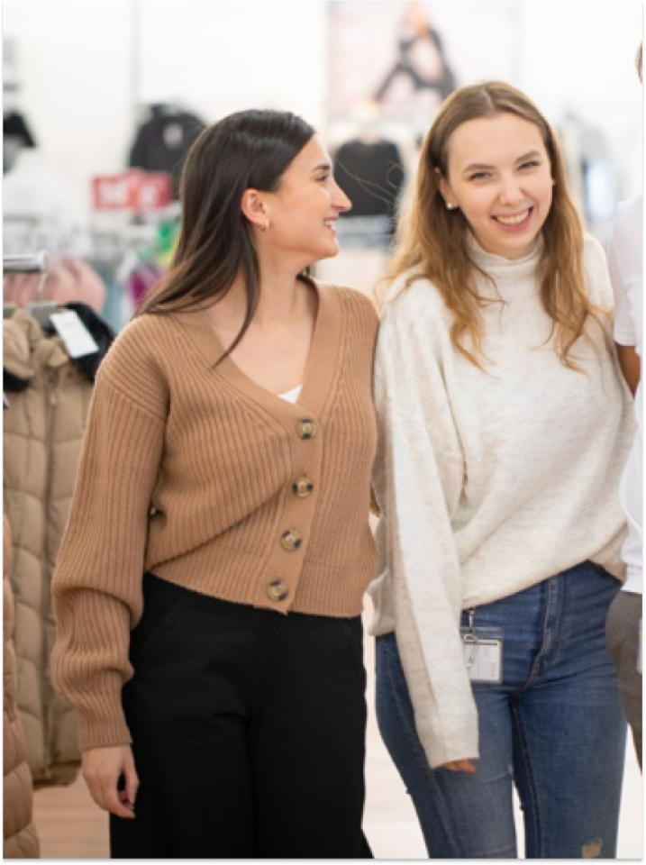 Zwei Frauen stehen drinnen, lächeln und schauen sich an. Die eine trägt eine hellbraune Strickjacke und eine schwarze Hose, die andere einen weißen Pullover und Jeans. Sie wirken glücklich und entspannt in einer ungezwungenen Umgebung, möglicherweise einem Geschäft oder einem öffentlichen Ort.