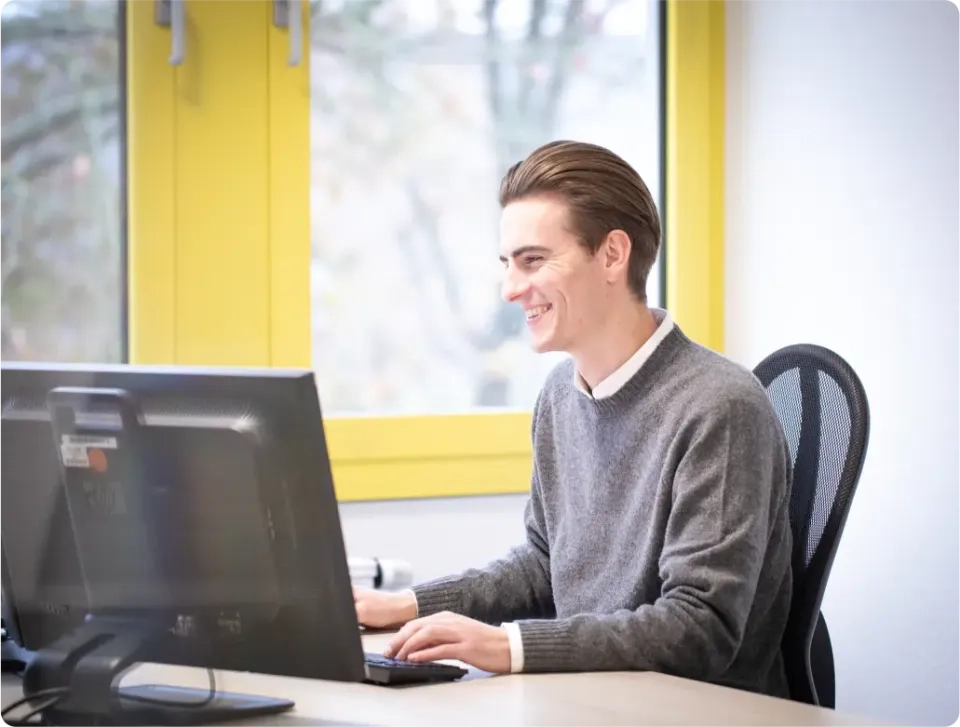 Ein junger Mann mit braunem Haar, einem grauen Pullover und einem weißen Hemd, sitzt an einem Schreibtisch und arbeitet am Computer. Er lächelt, im Hintergrund sind ein leuchtend gelber Fensterrahmen und verschwommene Bäume zu sehen.