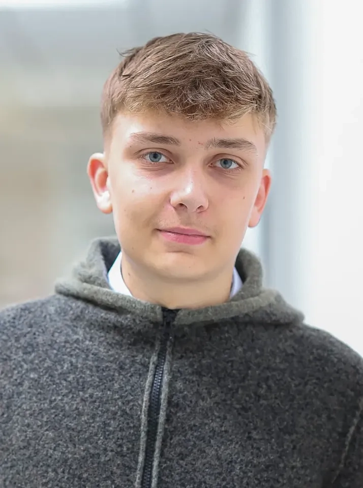 Ein junger Mann mit kurzen hellbraunen Haaren und blauen Augen trägt eine graue Fleecejacke mit Reißverschluss. Er steht in einem sanft beleuchteten Raum und blickt mit neutralem Gesichtsausdruck direkt in die Kamera.