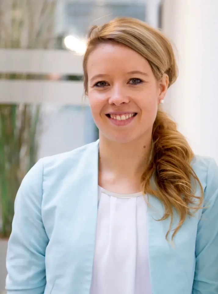 Eine Frau mit langen blonden Haaren, die zu einem tiefen Pferdeschwanz gebunden sind, trägt einen hellblauen Blazer über einem weißen Oberteil und lächelt in die Kamera, während sie in einem Innenbereich vor einem unscharfen Hintergrund steht.