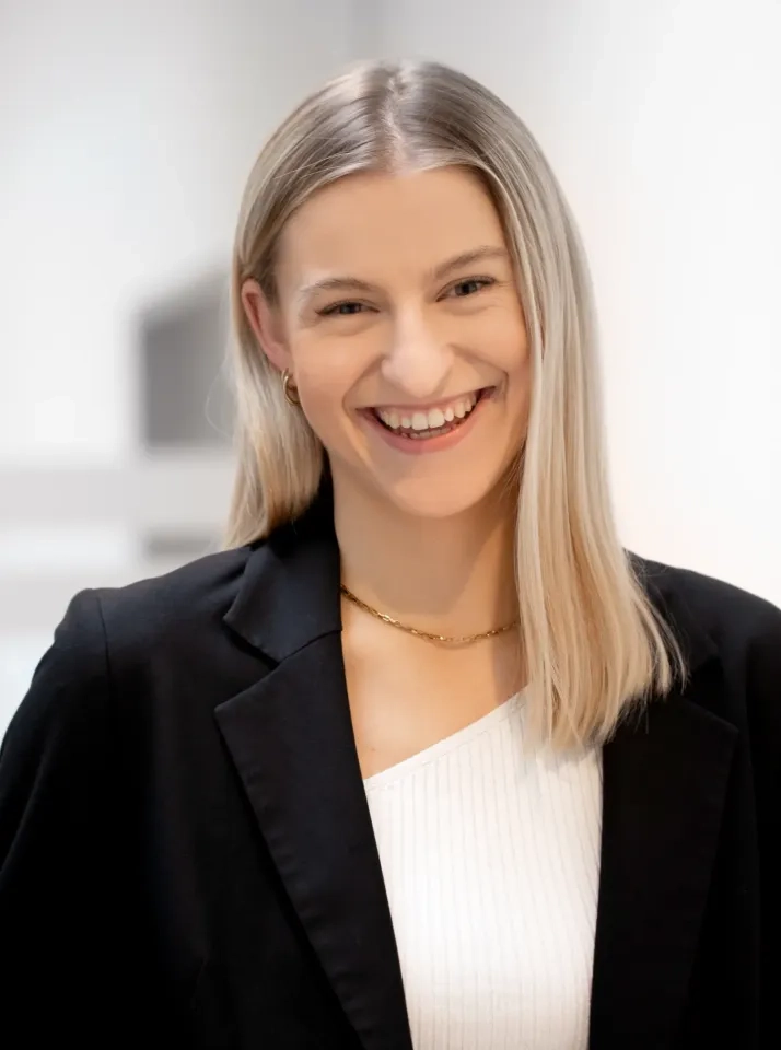 Eine Frau mit glattem blondem Haar, die einen schwarzen Blazer über einem weißen Oberteil und eine goldene Halskette trägt, lächelt in einem gut beleuchteten Innenbereich in die Kamera.