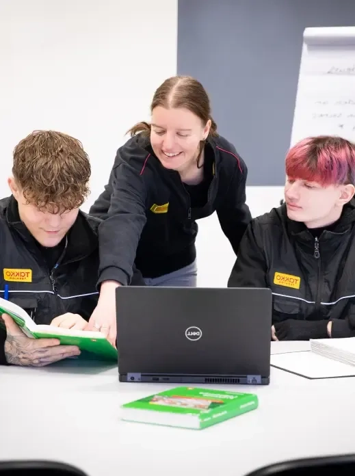 Eine Frau hilft zwei Schülern an einem Tisch mit Laptop und Büchern. Sie zeigt auf ein Buch, das einer der Schüler in der Hand hält, während der andere aufmerksam zuhört. Alle tragen schwarze Jacken mit gelben TACKID-Logos.