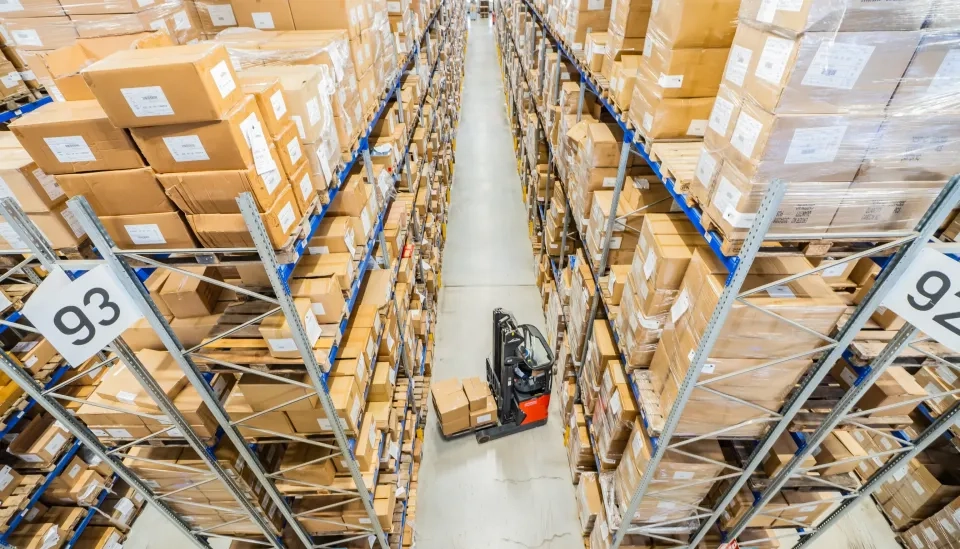 Ein Arbeiter bedient einen Gabelstapler in einem Lagergang, der mit hohen Regalen gesäumt ist, auf denen zahlreiche braune Kartons stapeln. Die Regale sind mit Nummern beschriftet, und die Szene ist von oben hell beleuchtet.
