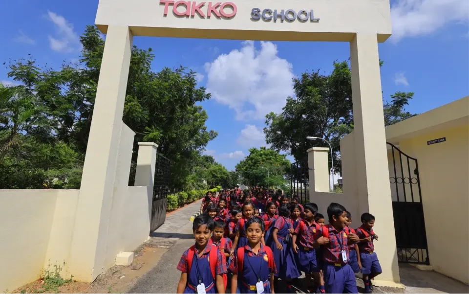 Eine große Gruppe von Kindern in passenden rot-blauen Schuluniformen verlässt an einem sonnigen Tag, umgeben von Bäumen und Grün, das Haupttor einer Schule mit dem Namen „Takko School“.