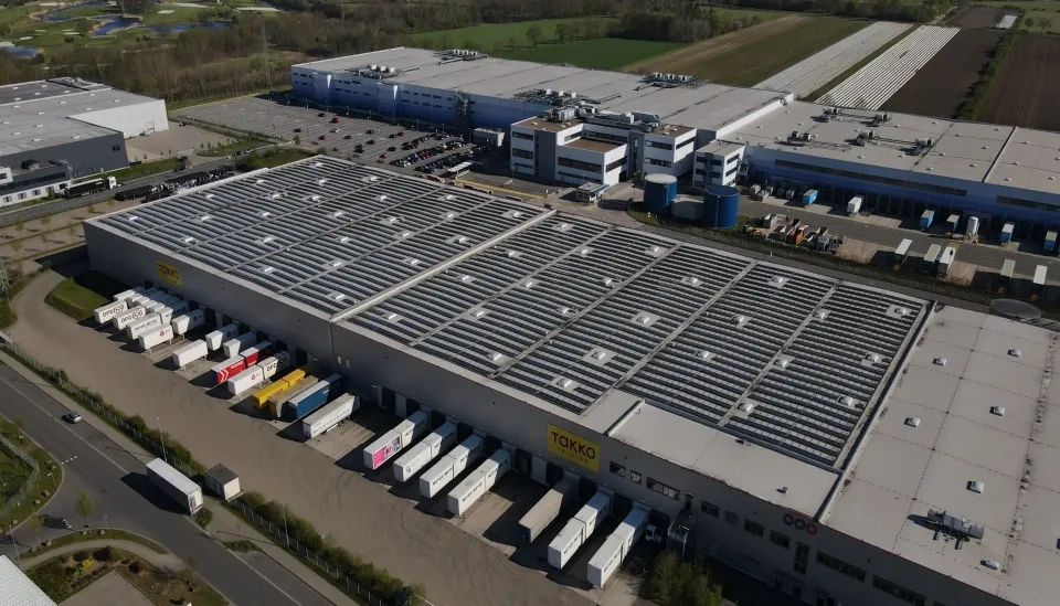 Luftaufnahme eines großen Lagerkomplexes mit Solarmodulen auf dem Dach. Draußen parken mehrere Laderampen mit LKWs. Das Gelände ist von Bürogebäuden, Parkplätzen und Grünflächen umgeben.