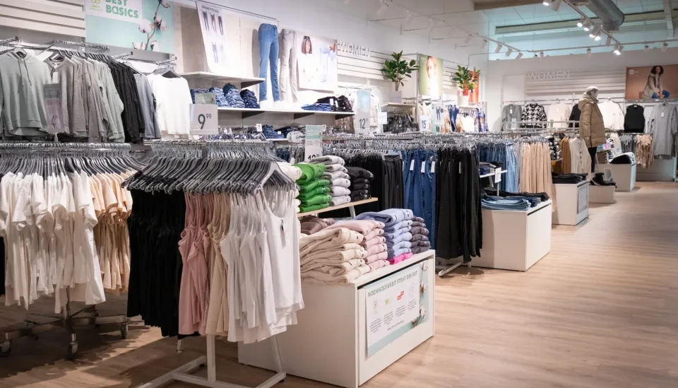 Ein ordentlich organisiertes Bekleidungsgeschäft mit Regalen voller Hemden, Hosen und Pullover. Gefaltete Kleidung liegt auf Tischen, und der helle Raum hat Holzböden und weiße Wände mit Modepostern und Pflanzen.