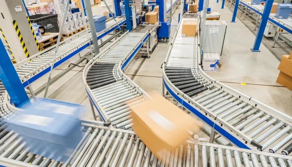 Pakete bewegen sich schnell entlang eines Systems aus Metallförderbändern in einem Lagerhaus oder Vertriebszentrum, umgeben von blauen Balken und verschiedenen Maschinen, wobei einige Kartons aufgrund der Bewegung unscharf sind.