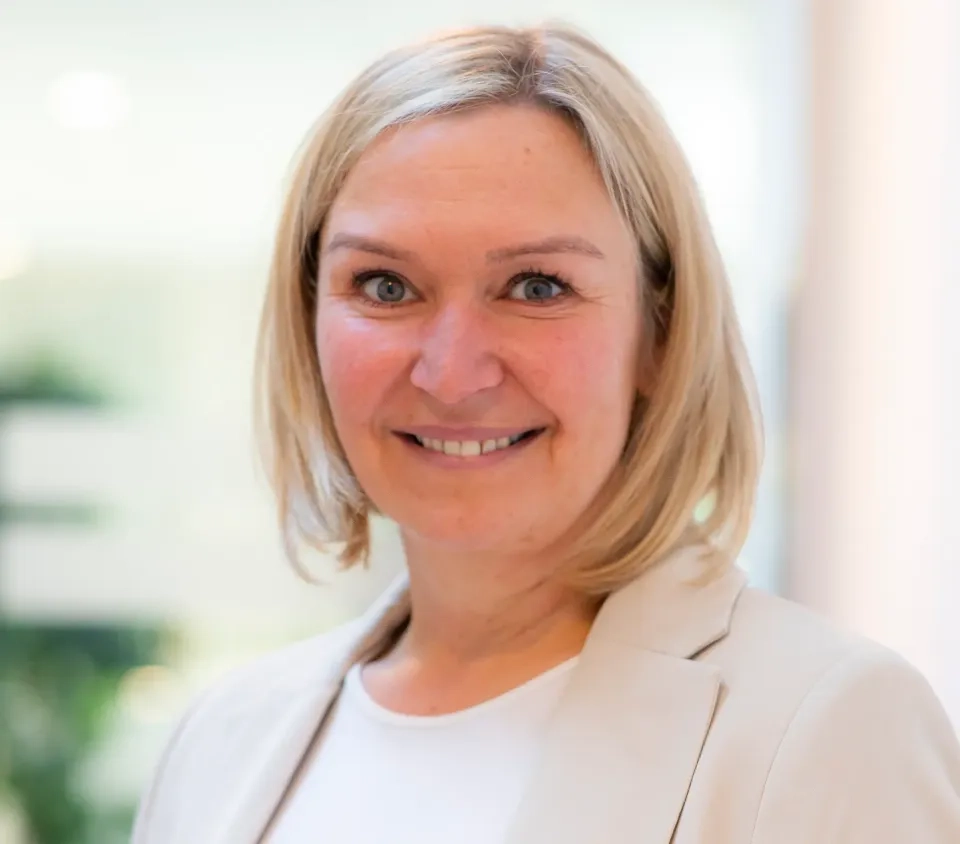 Eine Frau mit glattem blondem Haar, die einen hellen Blazer und ein weißes Oberteil trägt, lächelt in einer hellen, unscharfen Innenumgebung in die Kamera.