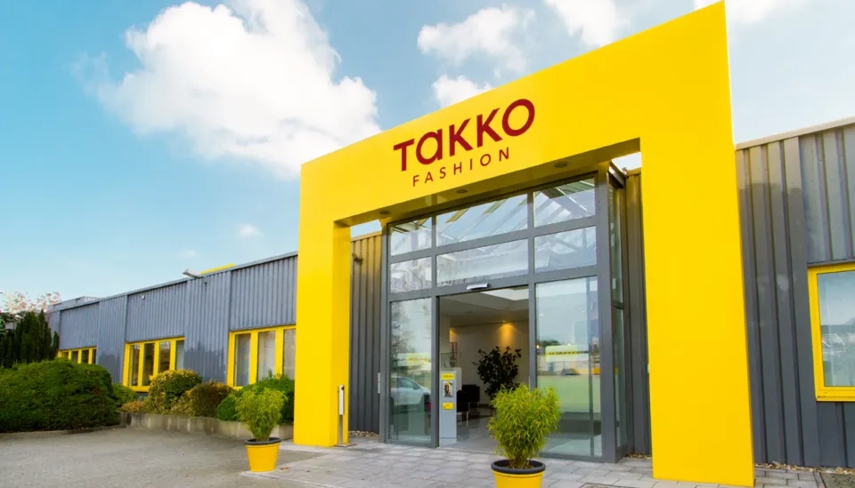 Leuchtend gelber Eingang eines modernen Takko Fashion Stores mit großen Glastüren, Topfpflanzen auf beiden Seiten und einem blauen Himmel mit Wolken im Hintergrund.