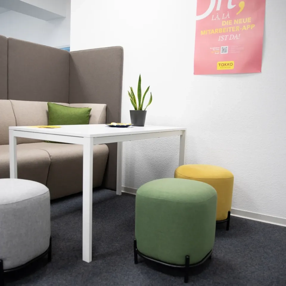 Eine moderne Bürolounge mit einem grauen Hochlehnersofa, einem weißen Tisch, einer Topfpflanze und drei runden Hockern in Grau, Grün und Gelb. An der weißen Wand über dem Tisch hängt ein rosa Poster.